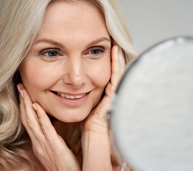 Happy 50s middle aged woman model touching face skin looking in mirror. Smiling mature older lady pampering, enjoying healthy skin care, aging beauty, skincare treatment cosmetic products concept.
